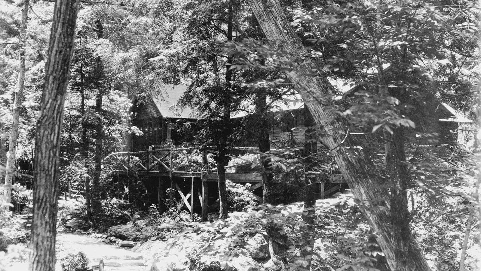 A black and white photograph of a rustic log cabin surrounded by forest.
