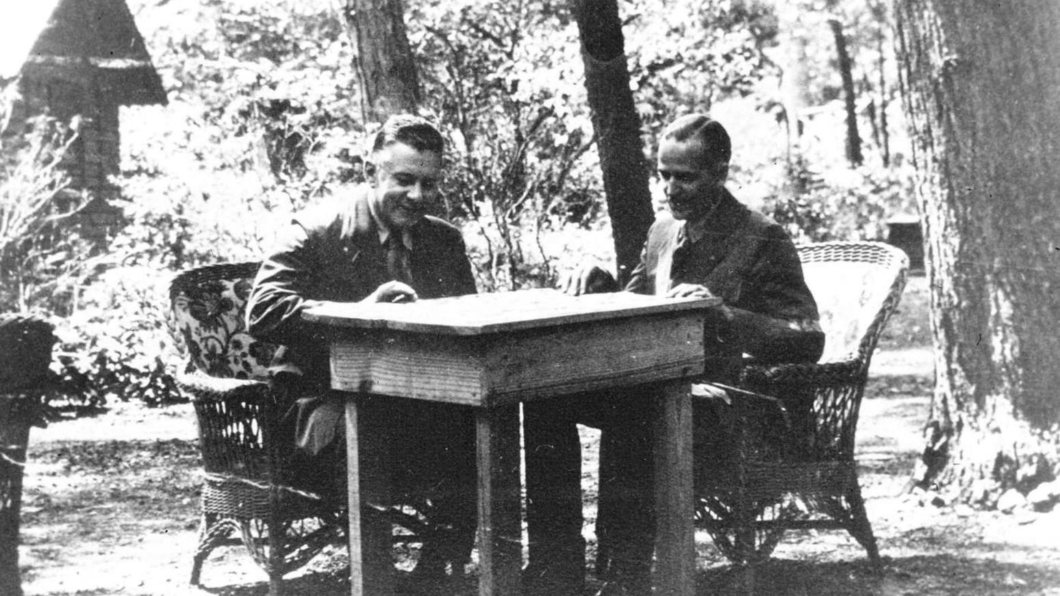 Two men sit at a table outdoors working a jigsaw puzzle.