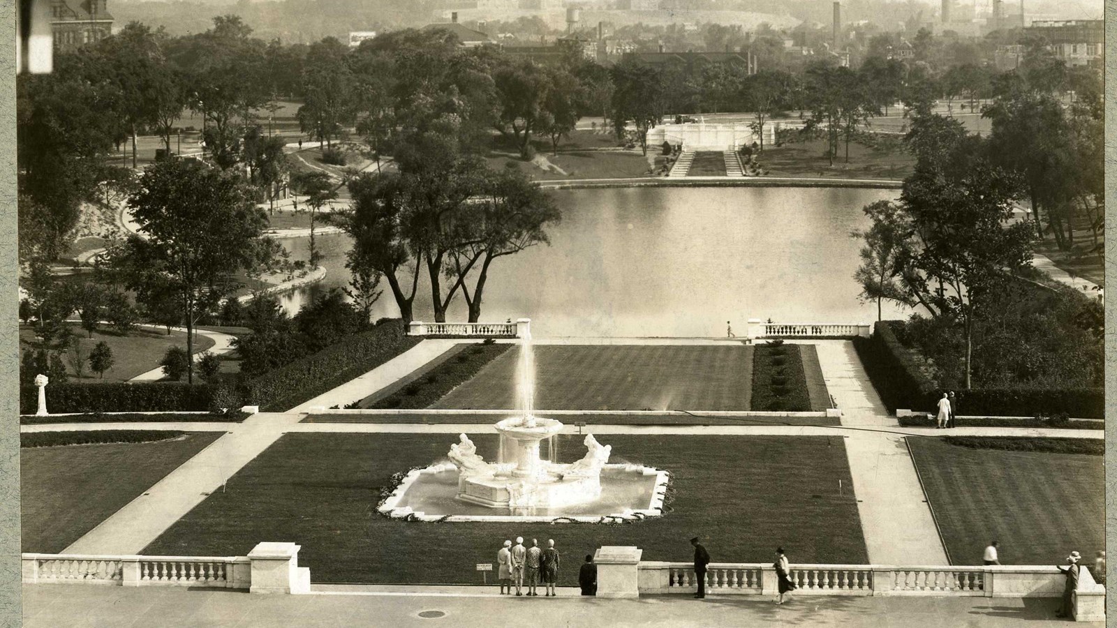 Black and white of grassy hill with fountain on it lined with trees leading to large body of water. 