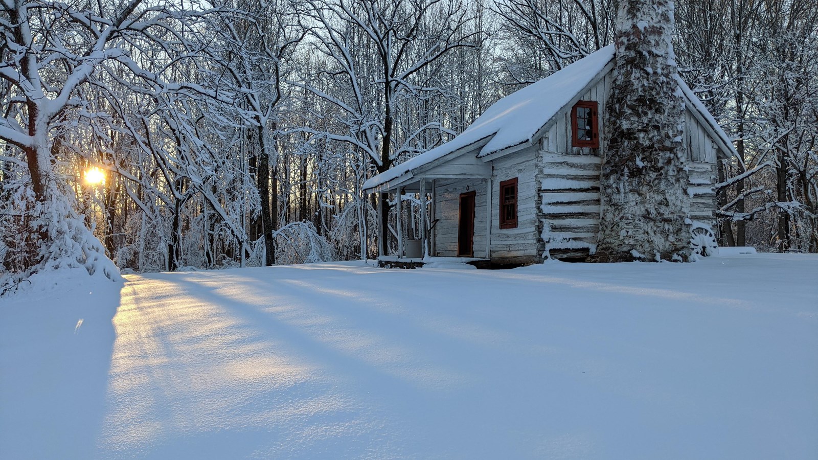 A log cabin with red window frames and a stone chimney stands in the midst of a snowy field.