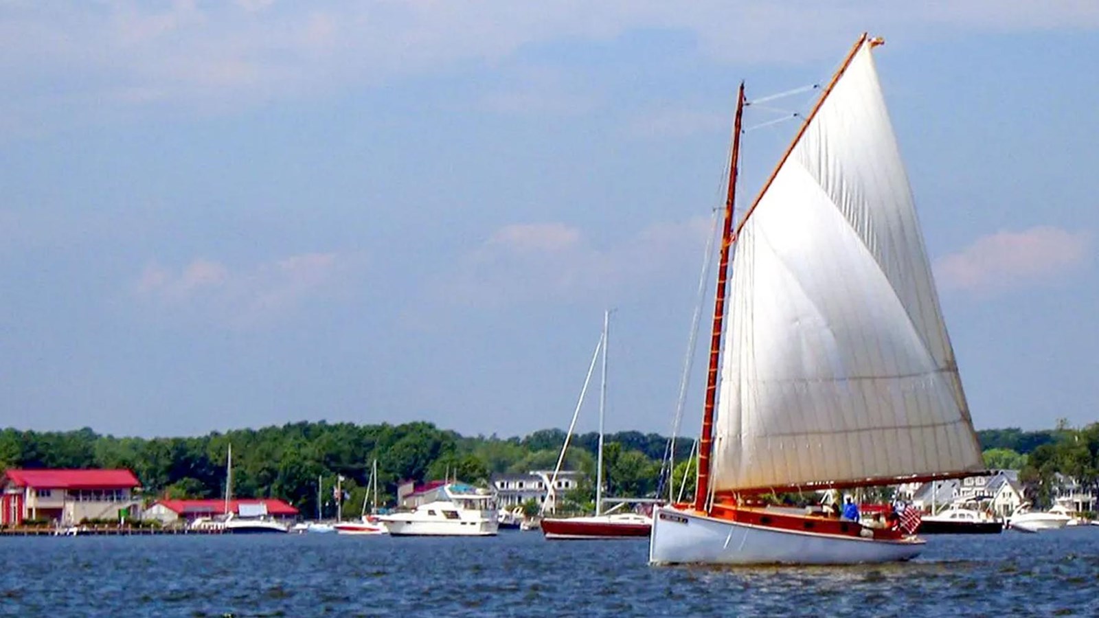 A sailboat with a large white sail on a calm body of water near a marina, under a partly cloudy sky.