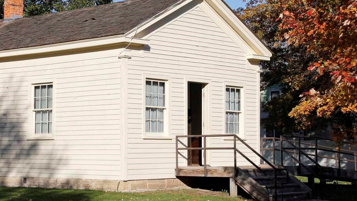 A one-story wooden building painted off-white has two windows and a central doorway.