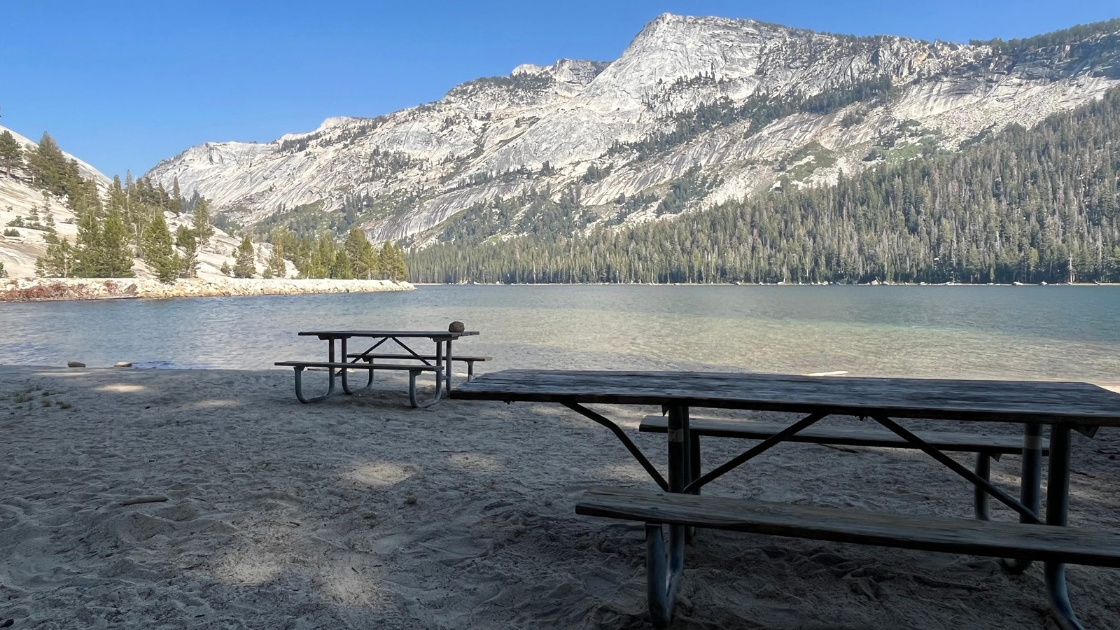 Two picnic tables on a sandy beach with a large lake and towering peaks