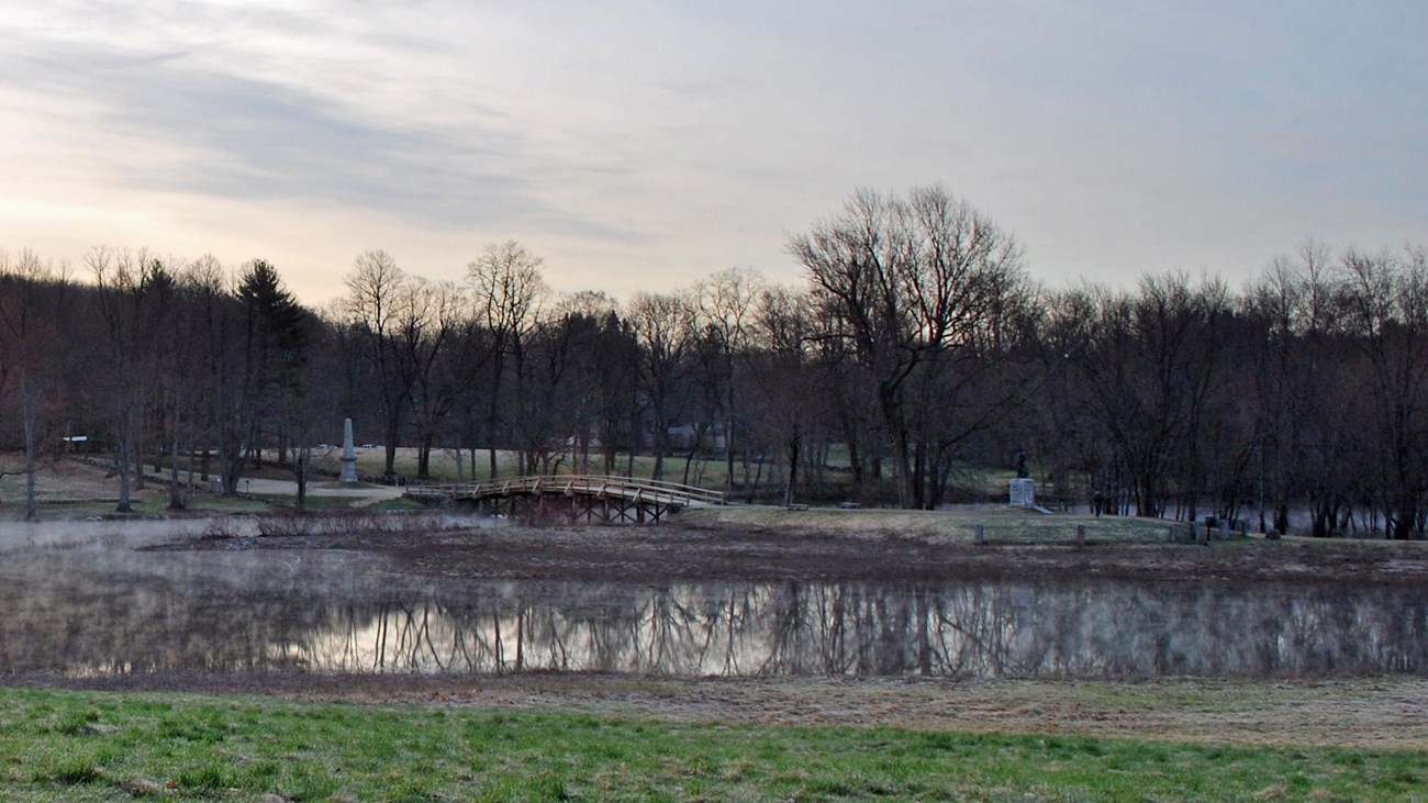 A wooden bridge spans a small river at sunrise. Water is flooded from the river surrounding