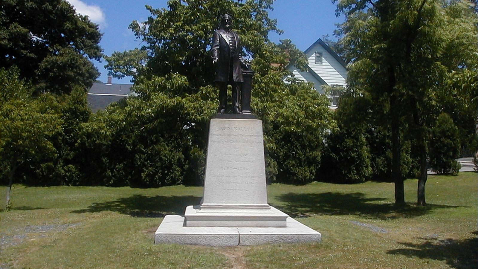 Statue of Adin Ballou sits on a pedestal in a grassy area surrounded by trees