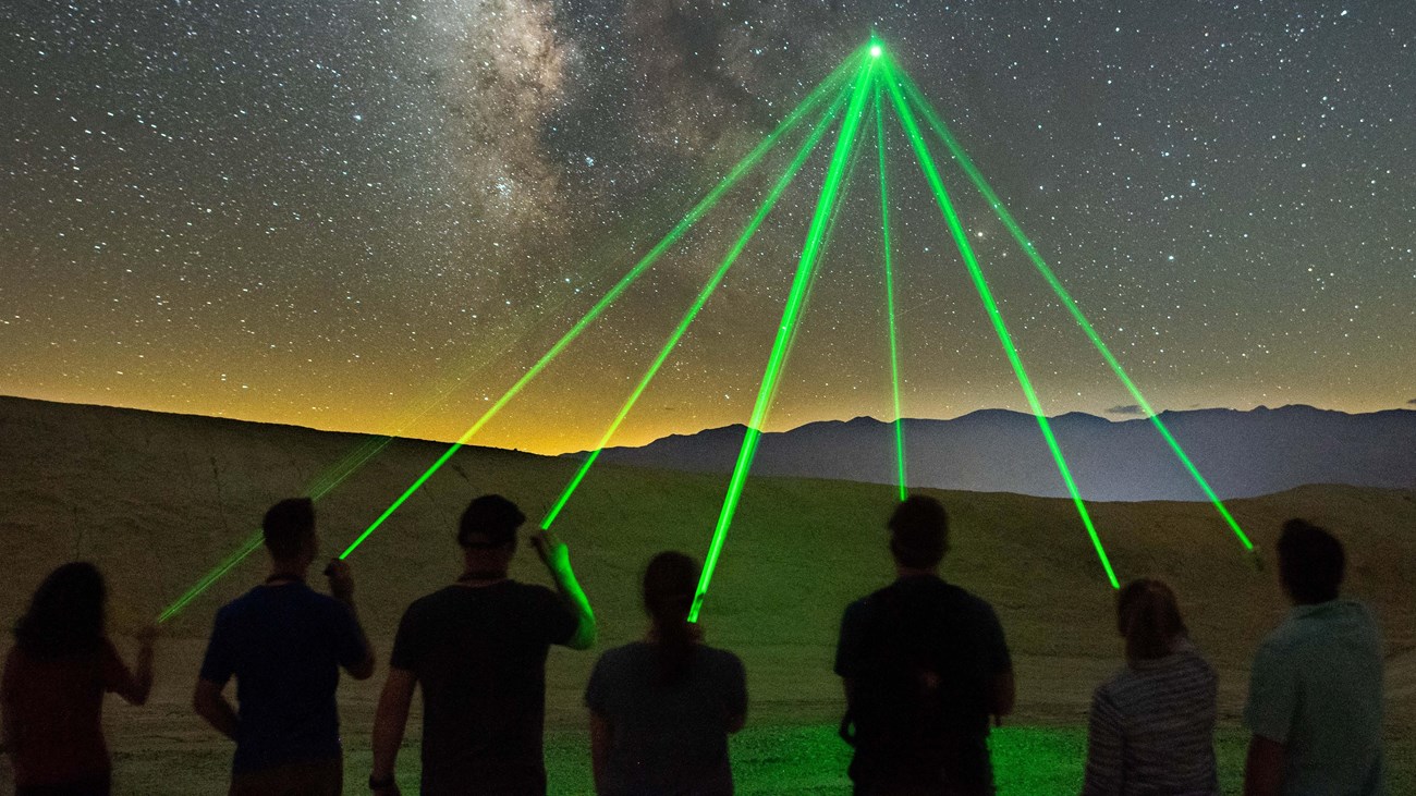 Seven people silhouetted pointing green lasers to the same spot in a dark night sky. 