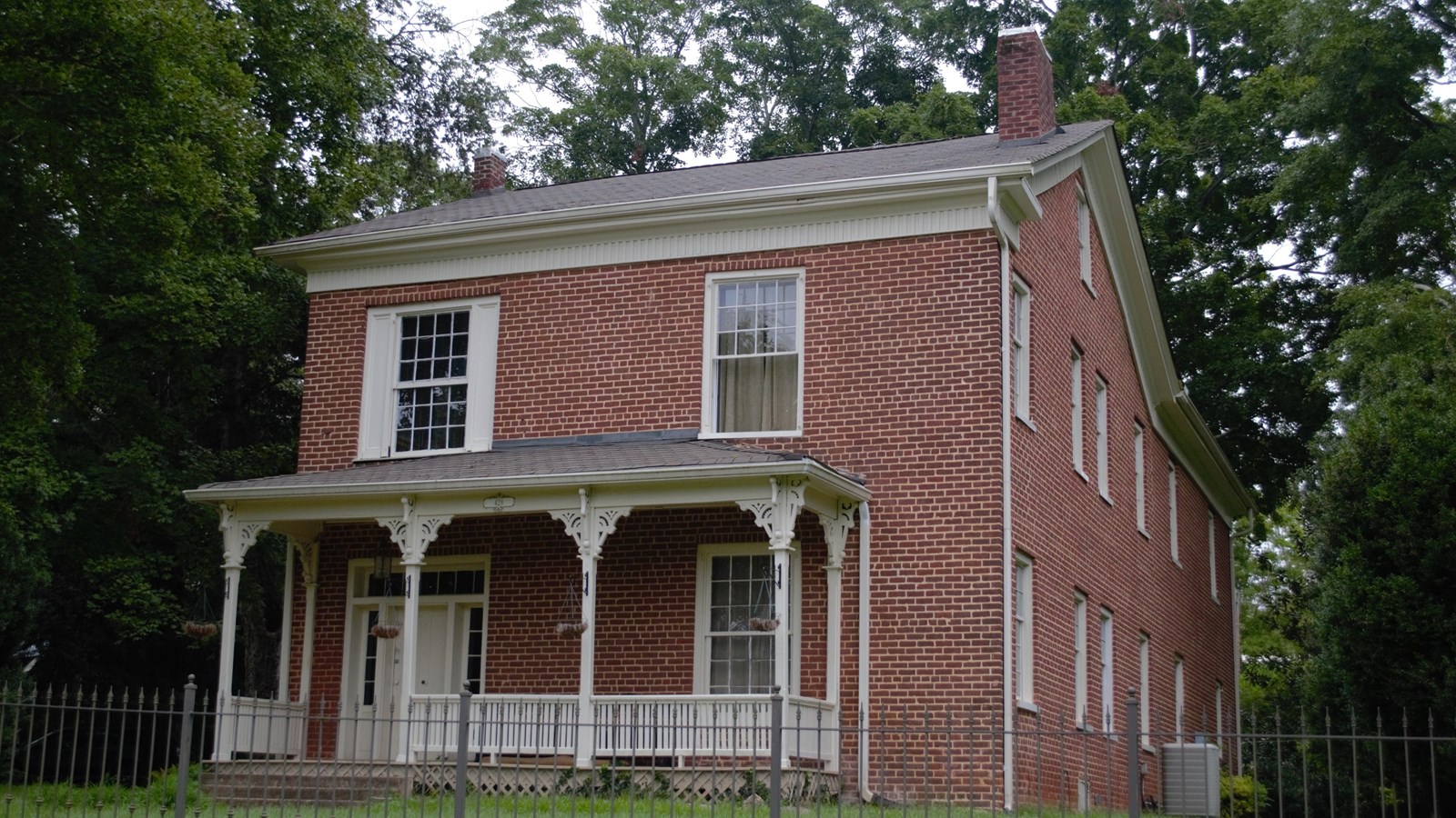 A two-story red brick house with white trims and columns. A wrought-iron fence surrounds the house.