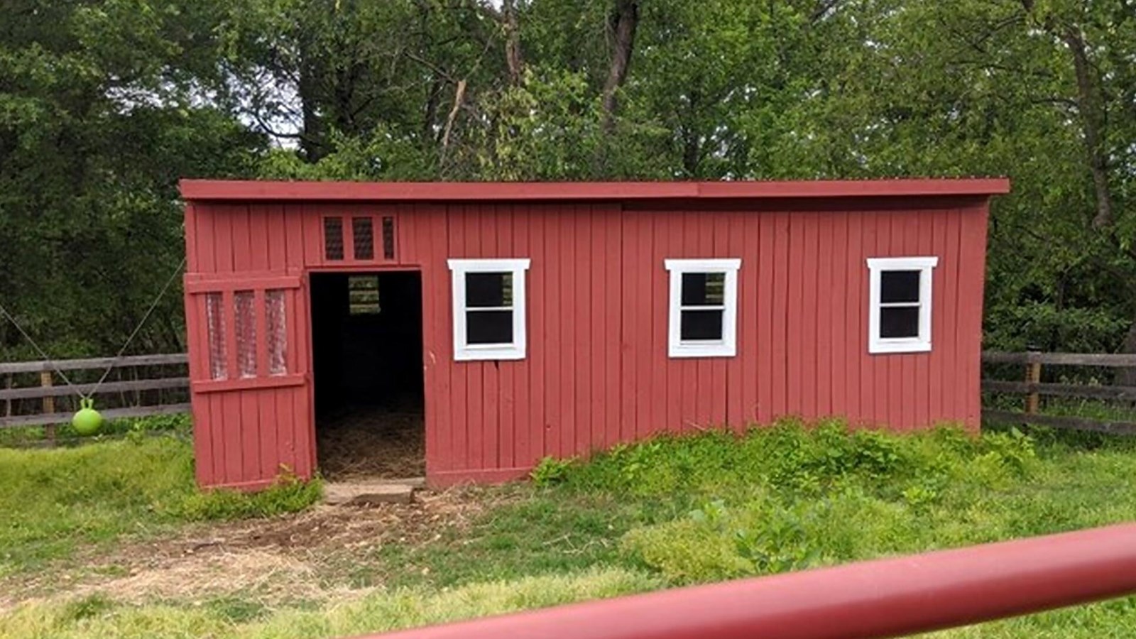 Photo of a red wooden building with white trimmed small windows.  Grass area around the pen.