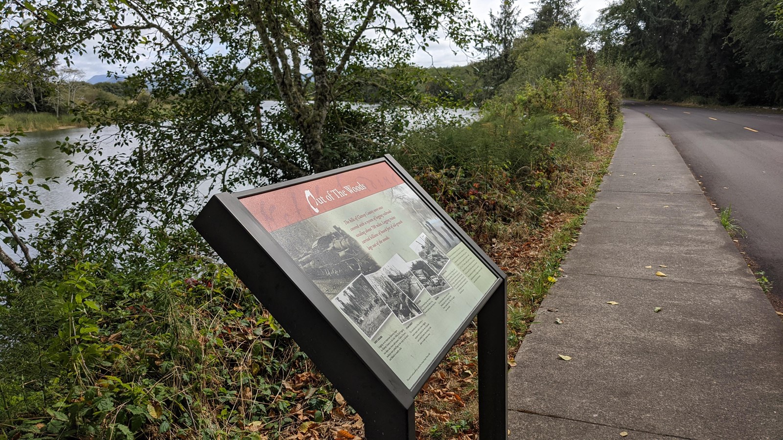 The Out of the Woods sign right alongside a sidewalk with the river behind it and the road in front