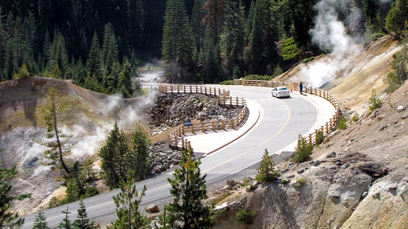 A road lined by wooden guardrails curves through a hydrothermal area of barren, steaming ground.