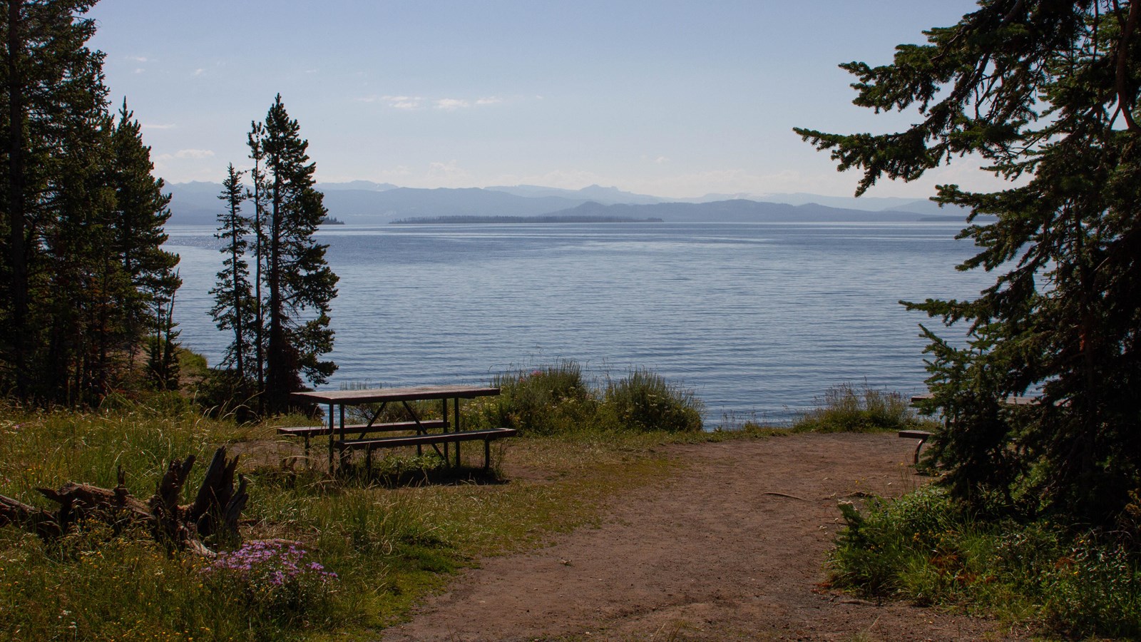 A few picnic tables are scattered by the shore of a large lake with distant mountains in background.