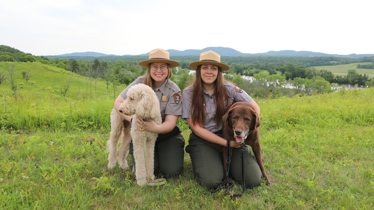 Two women in park ranger uniforms kneel next to two dogs on leash.