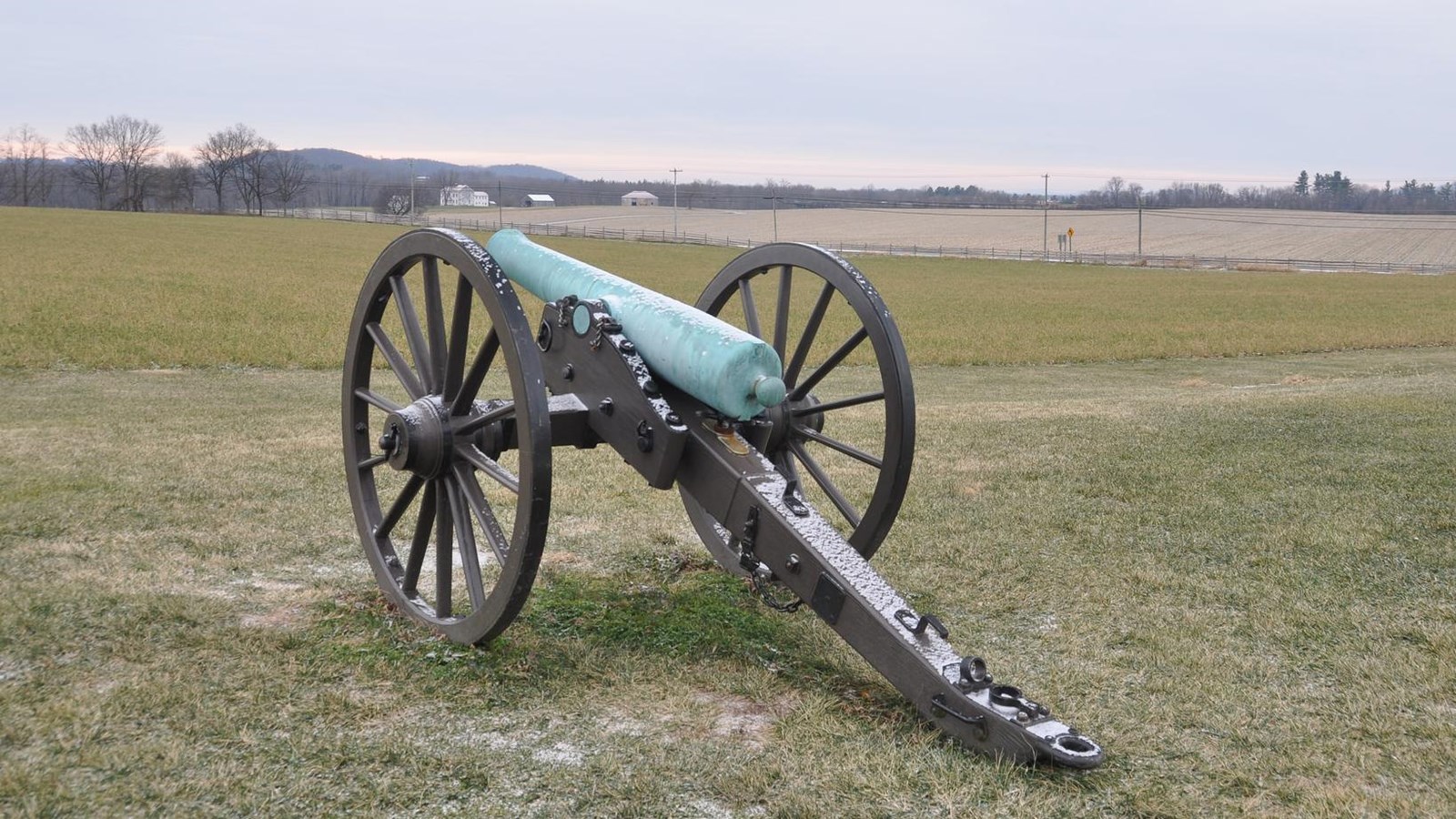 A cannon sits in a grassy field lightly dotted with snow.
