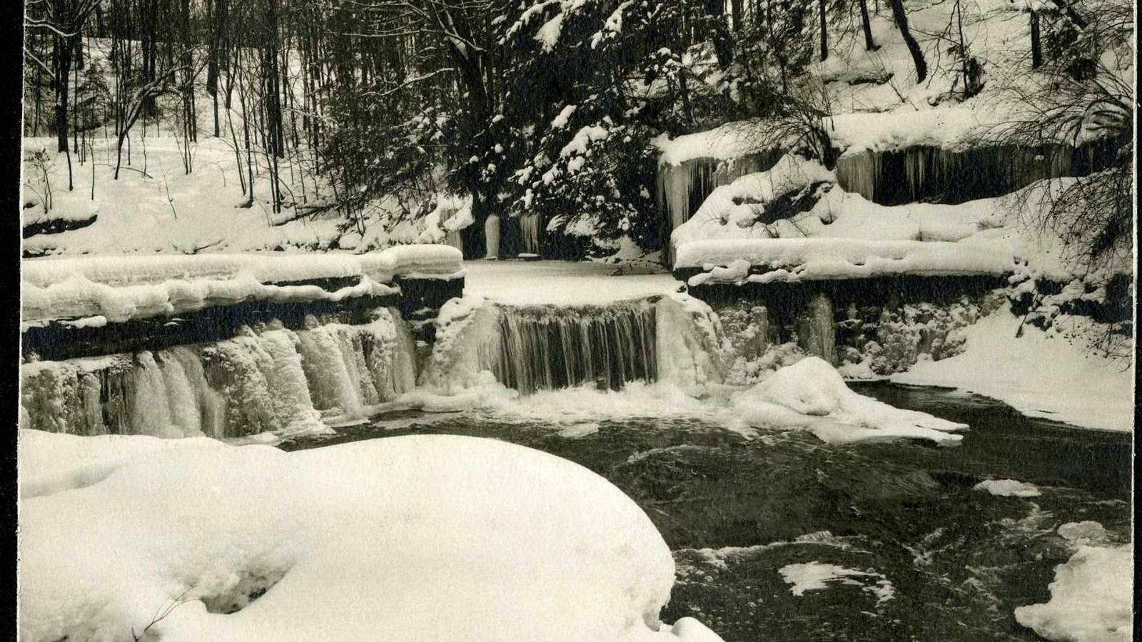 Black and white of river going down a small ledge with snow on the ground and trees in the back