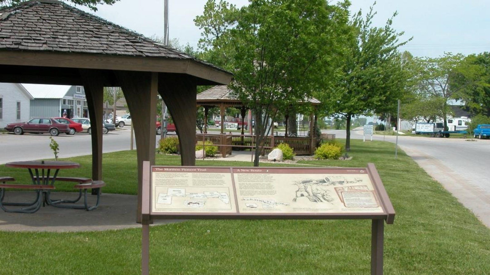 An interpretive exhibit panel sits in a grassy park with covered picnic tables.