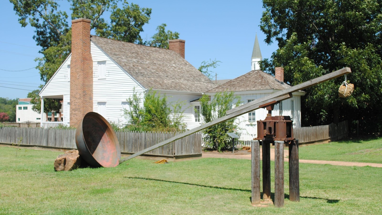A white building surrounded by a wooden fence. A wooden and metal structure sits in front.