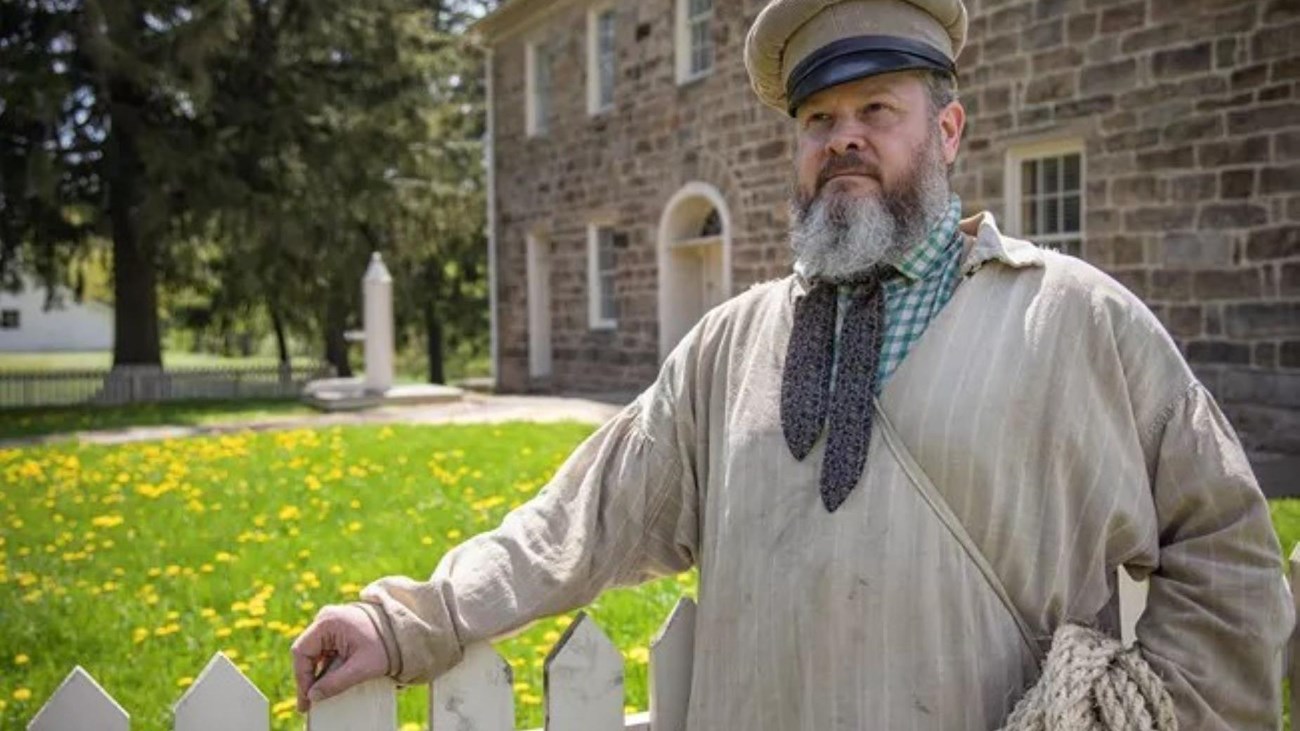 A man in period clothing holds rope in front of a stone tavern.