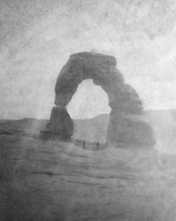 A black and white image of a large natural stone arch sitting alone on a cliff. 