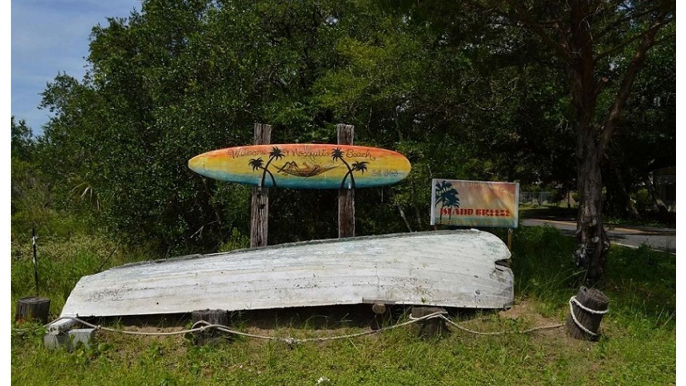 a turned over boat surrounded by grass with a painted surfboard stating “welcome to mosquito beach