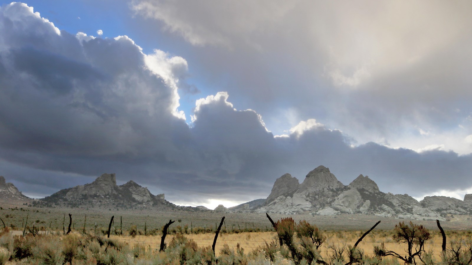 Stormy scene with sage brush, old fence, golden grass, and granite pinnacles in the backdrop.