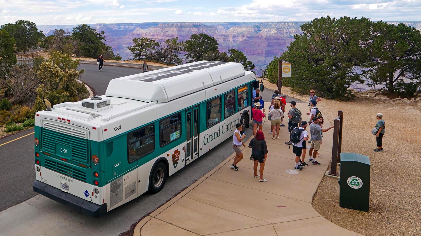a green and white bus with passengers at a bus stop with a vast canyon landscape in the distance.