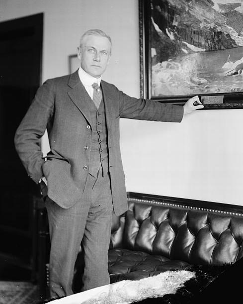 Stephen Mather, wearing a dark suit, stands in an office in front of a couch and painting.