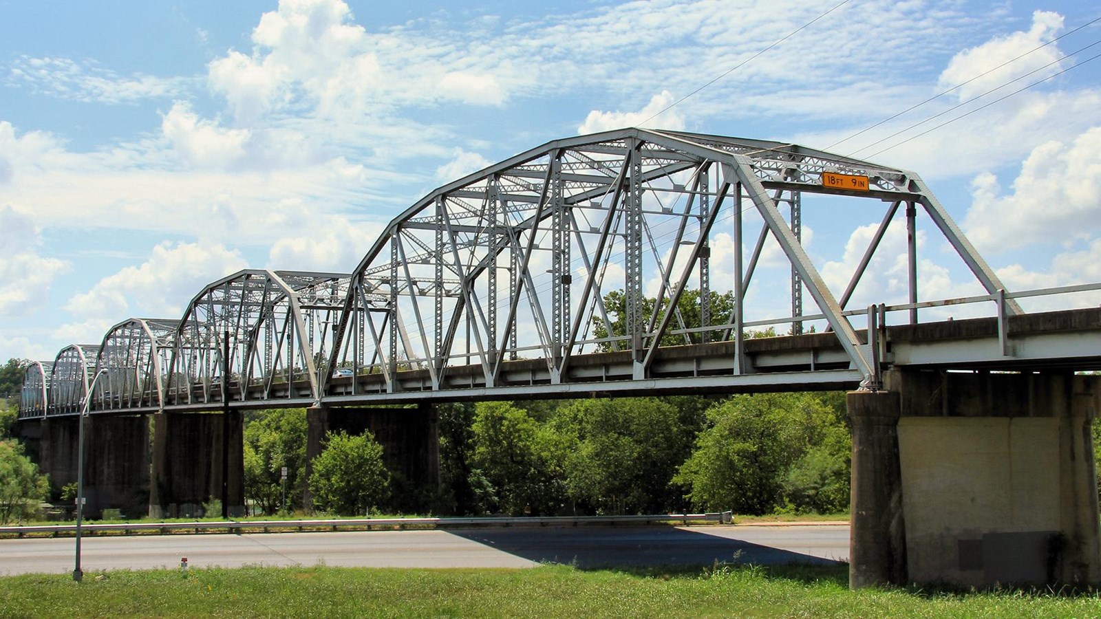 An elevated, steel bridge with trees and a highway beneath it