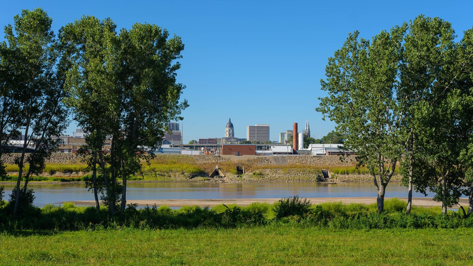 A channel of brown water is between small trees. The state capitol is in the distance.