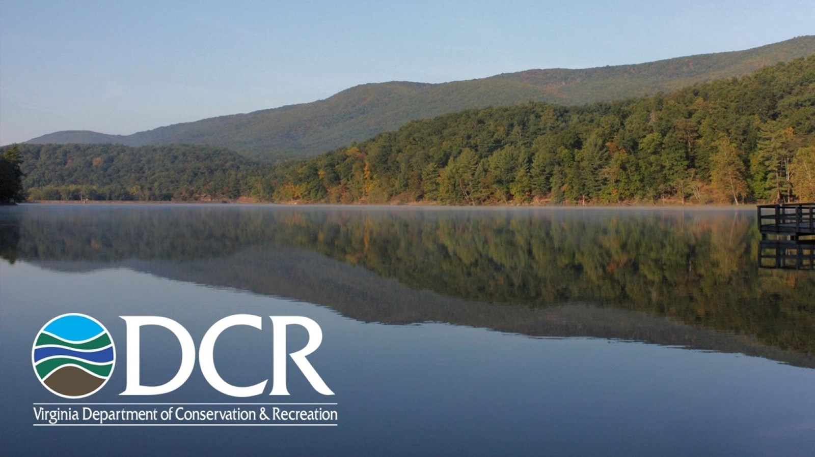 Misty mountain lake reflecting forested hills, autumn trees, and a wooden dock.