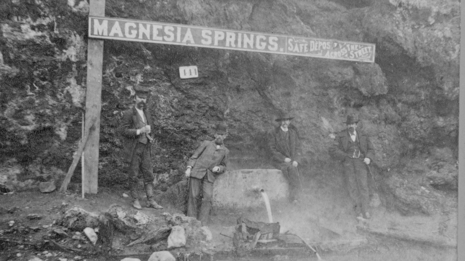 Four men next to large box with water pouring from a spout. Sign above them says 