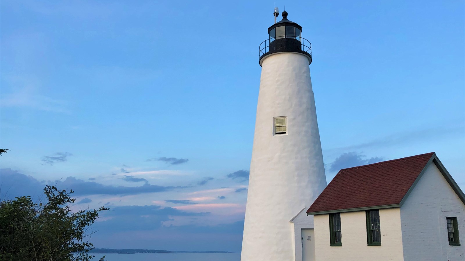 White lighthouse 100 feet tall with small white building attached under blue sky.