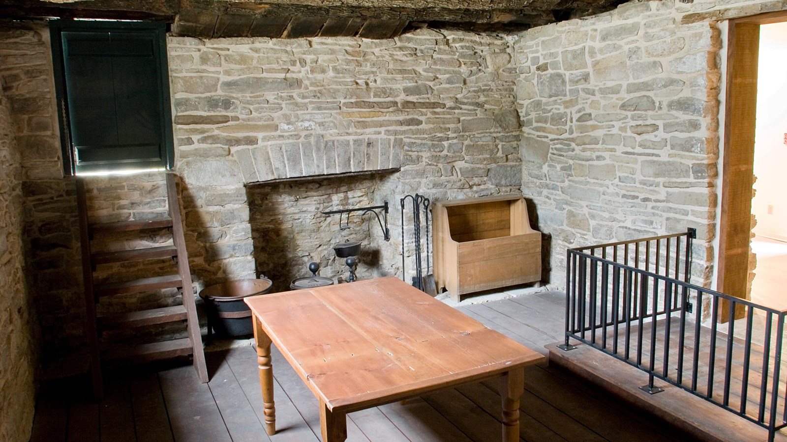 A brown table stands in front of a fireplace, in a stone basement. A small door is on the left.
