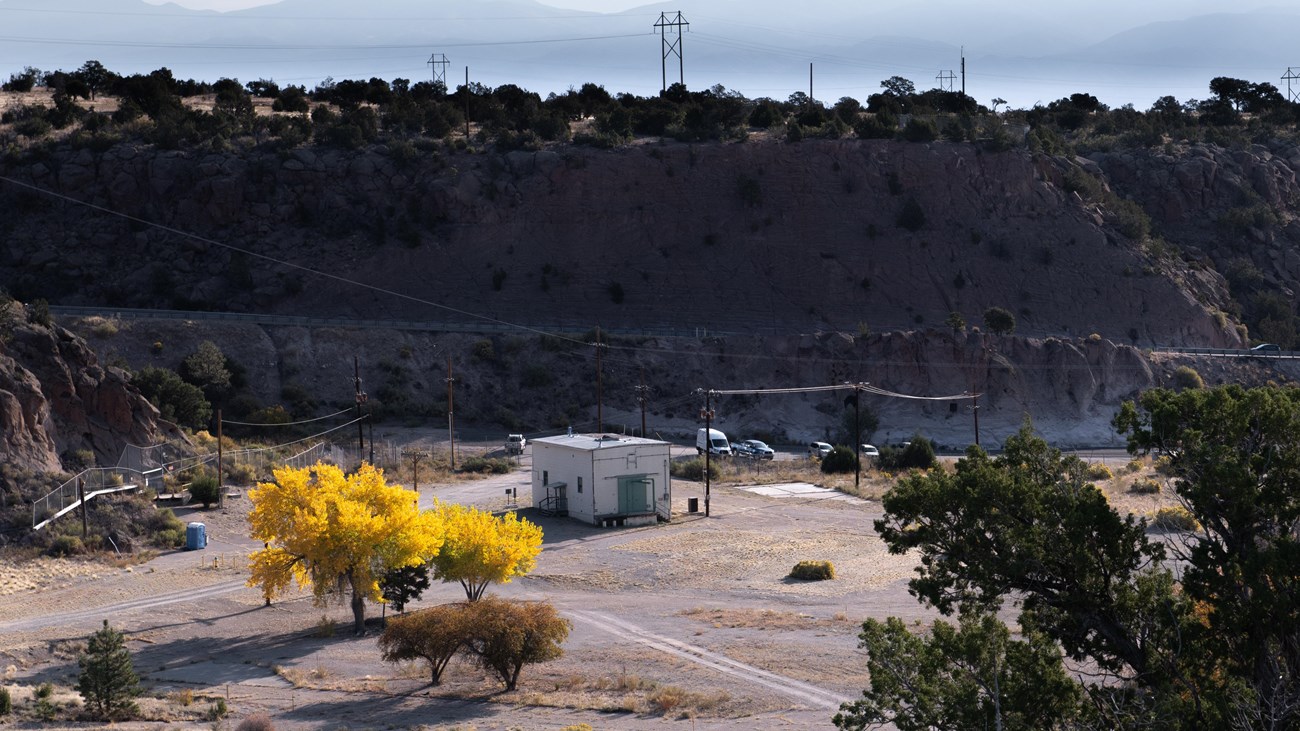 A small white building sits on a concrete pad in a valley surrounded by cliffs and sagebrush hills. 