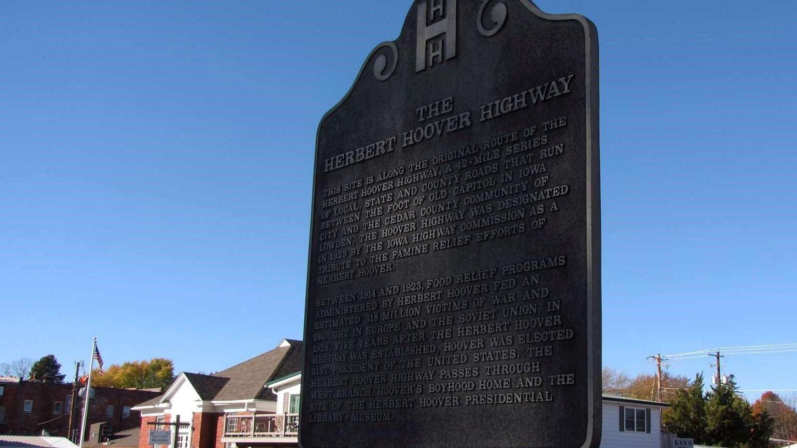 A bronze sign on a post marks the city street behind it as part of historic scenic highway.