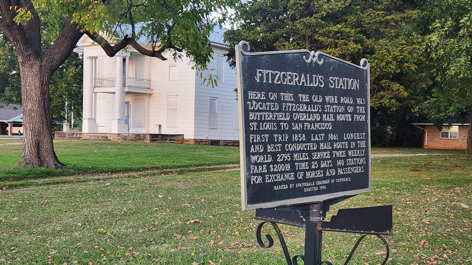 A blue sign sits on the lawn in front of a large white colonial house.