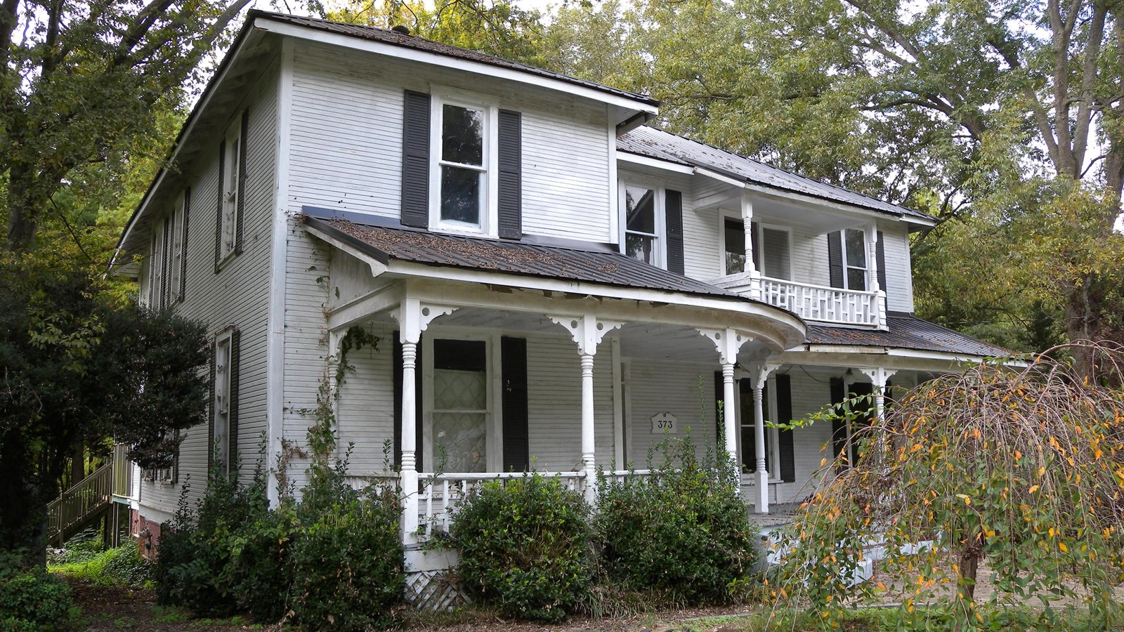 A two-story white colored house with a porch with decorative columns in a wooded area.