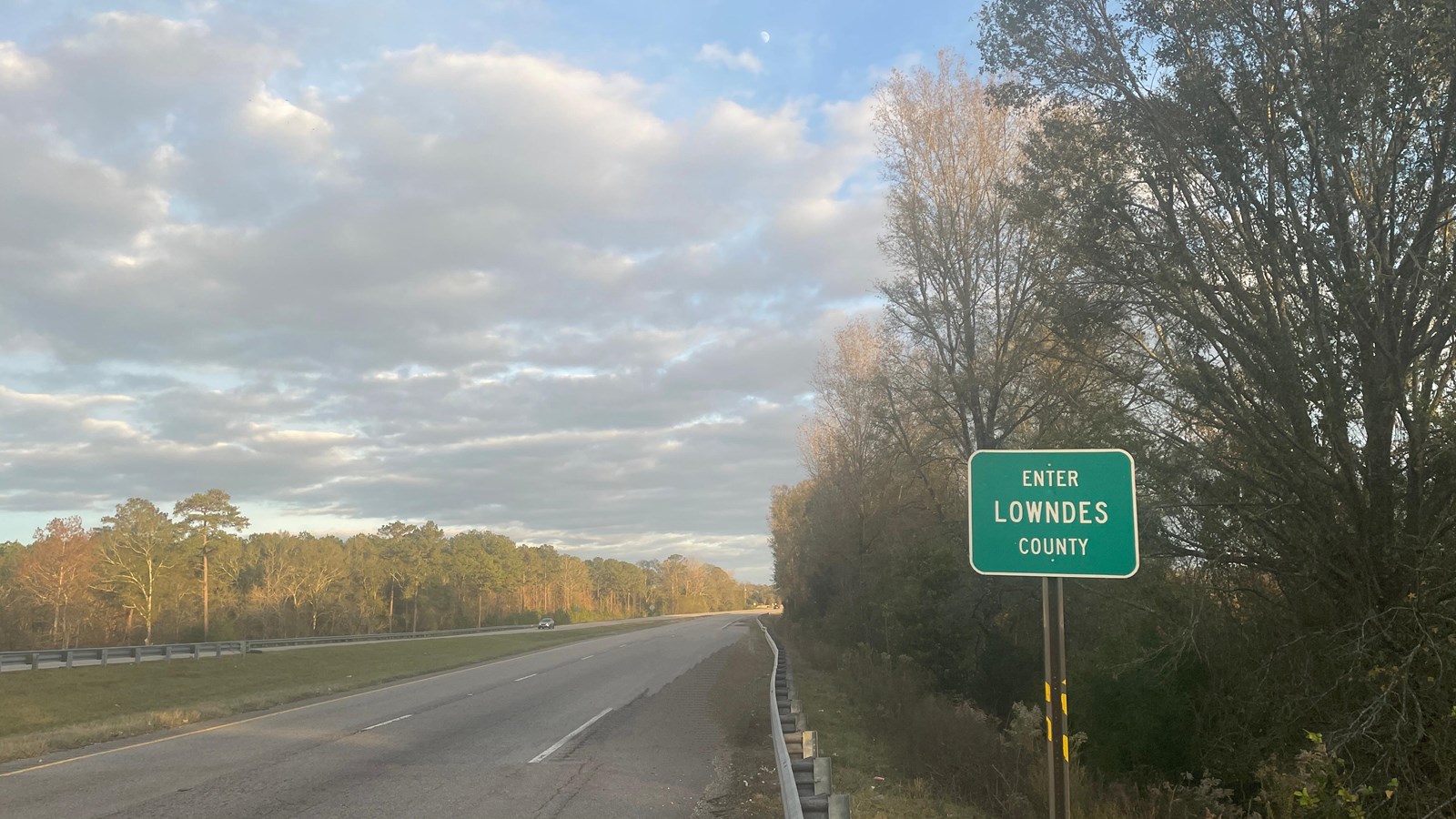 A green sign on the side of a two-lane highway reads, 