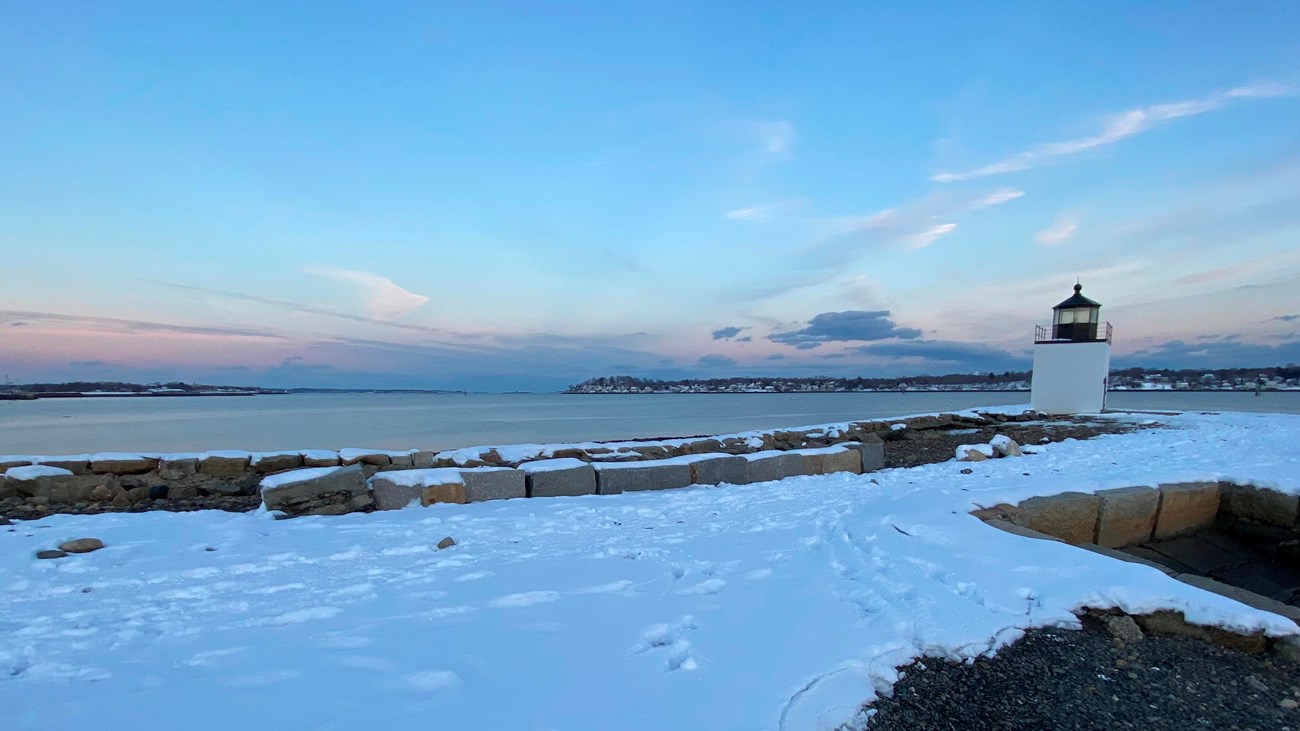 Light blue morning light over snowy wharf pathway with white lighthouse in distance.