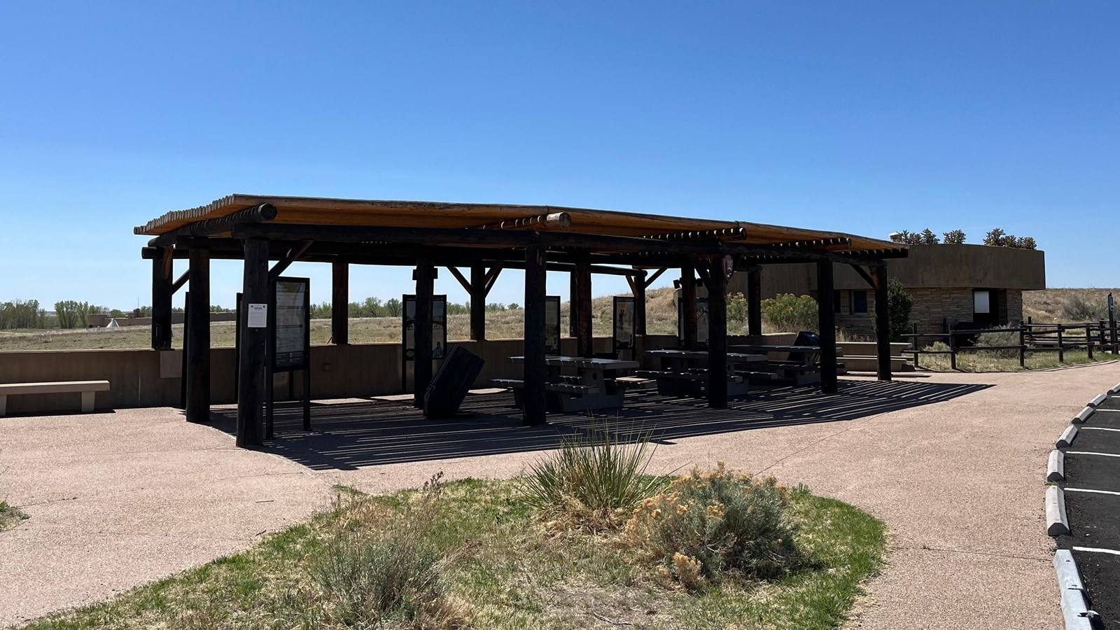 A wooden shelter with exhibits and picnic tables near a restroom and parking area.