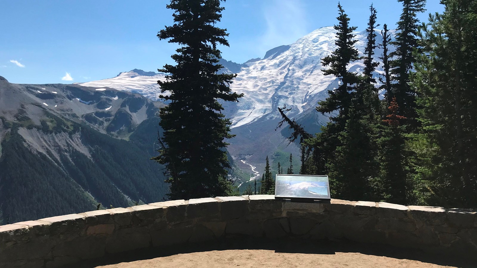 A wayside panel attached to a low rock wall overlooks a deep glacier valley and glaciated mountain.