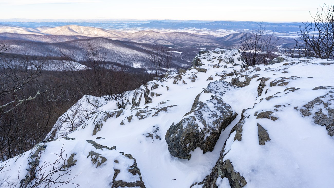 Snow covered rocks lie in the foreground of a view into a blue valley below.