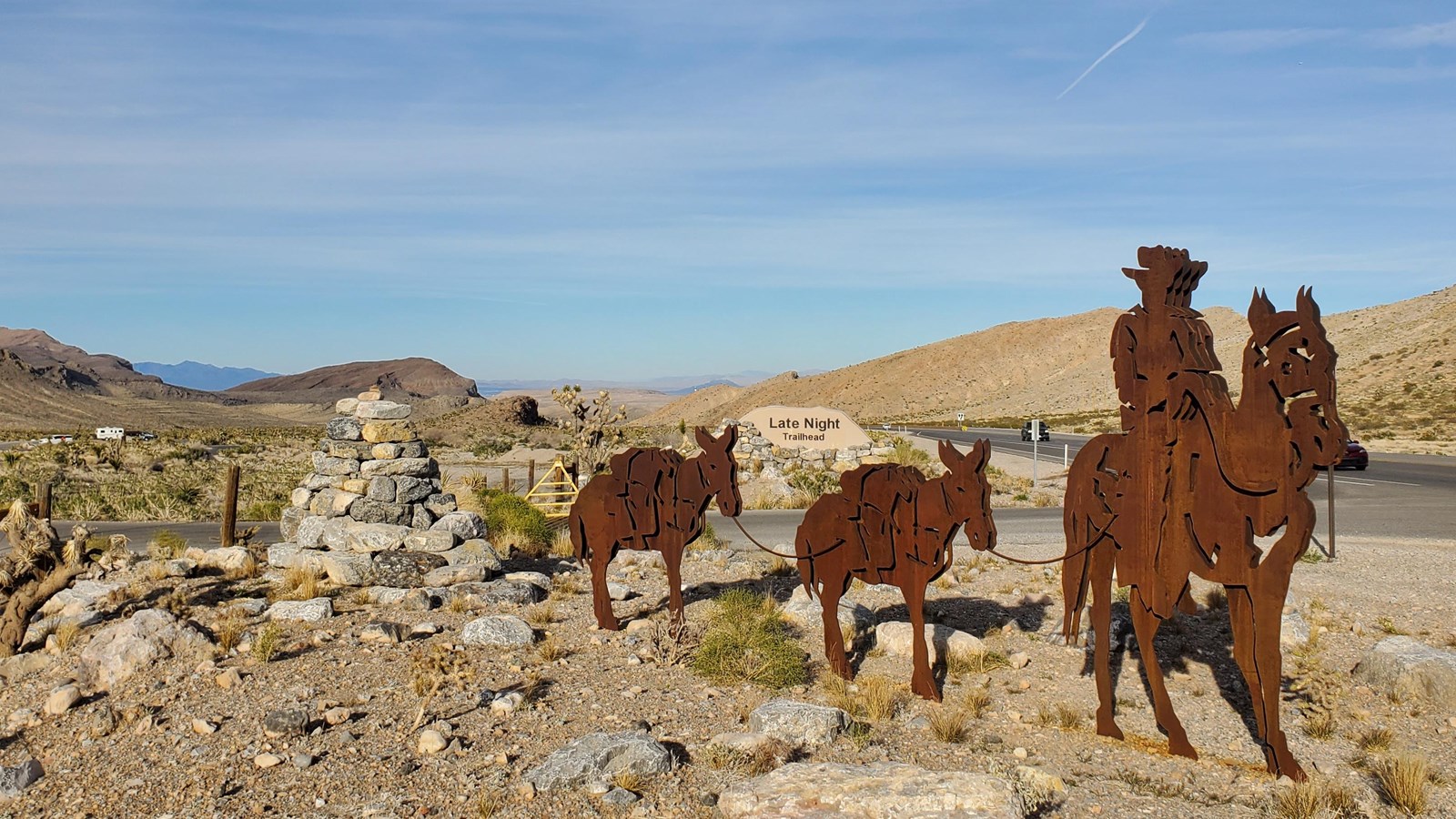 Metal silhouettes of rider on horseback followed by two mules, along roadway with sign reading 