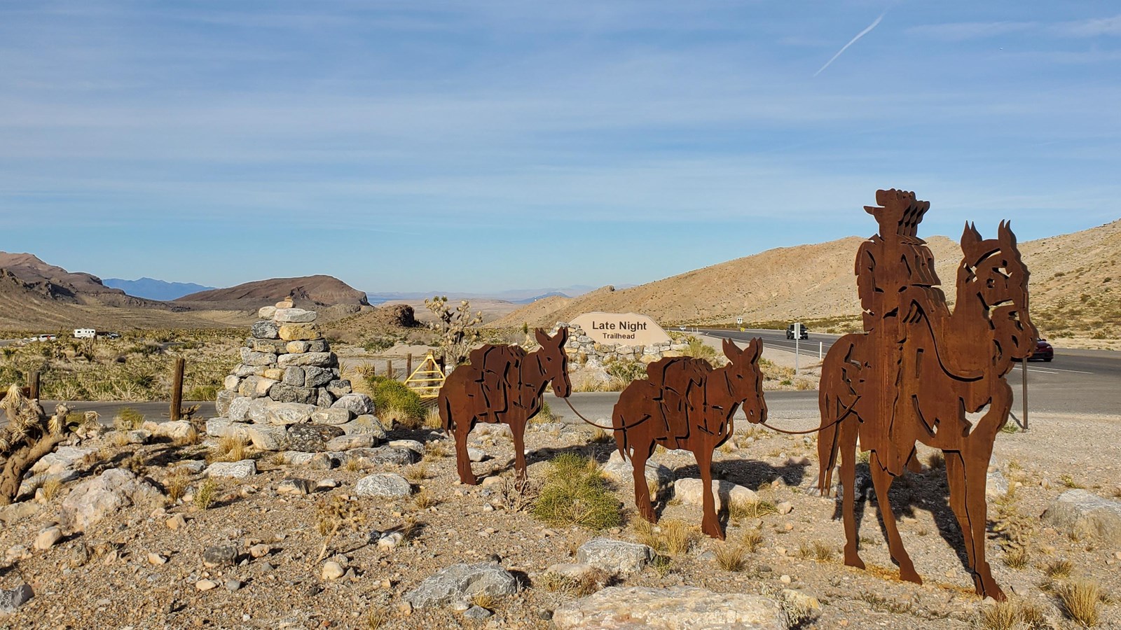 Metal silhouettes of rider on horseback followed by two mules, along roadway with sign reading