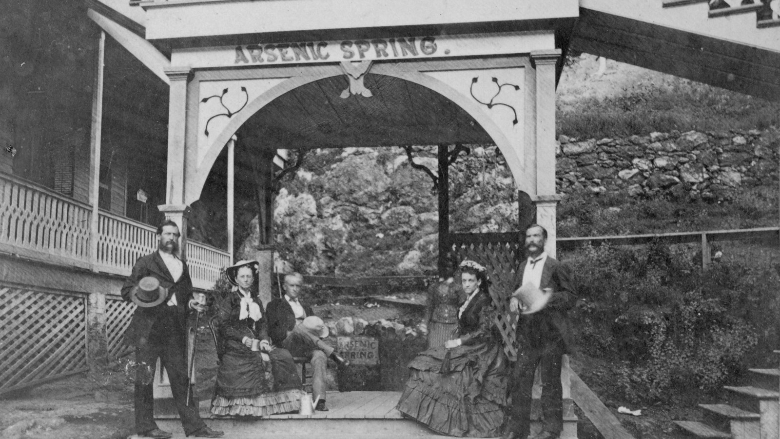Six well-dressed men & women in front of carved railings of Arsenic Spring covered porch.