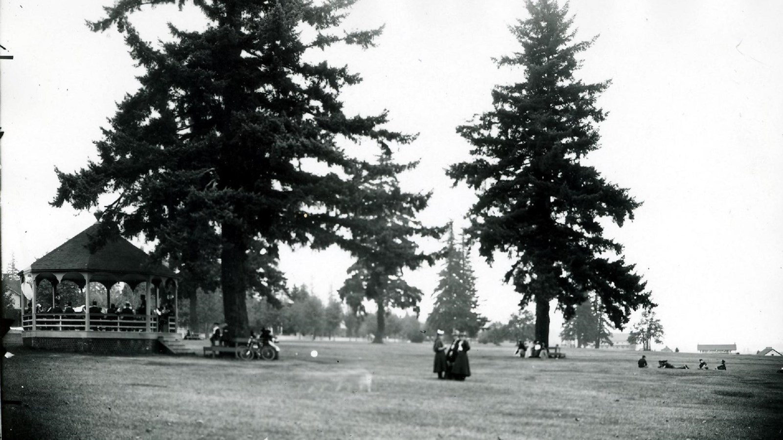 Black and white photo of a grassy field with trees, a bandstand, and people.