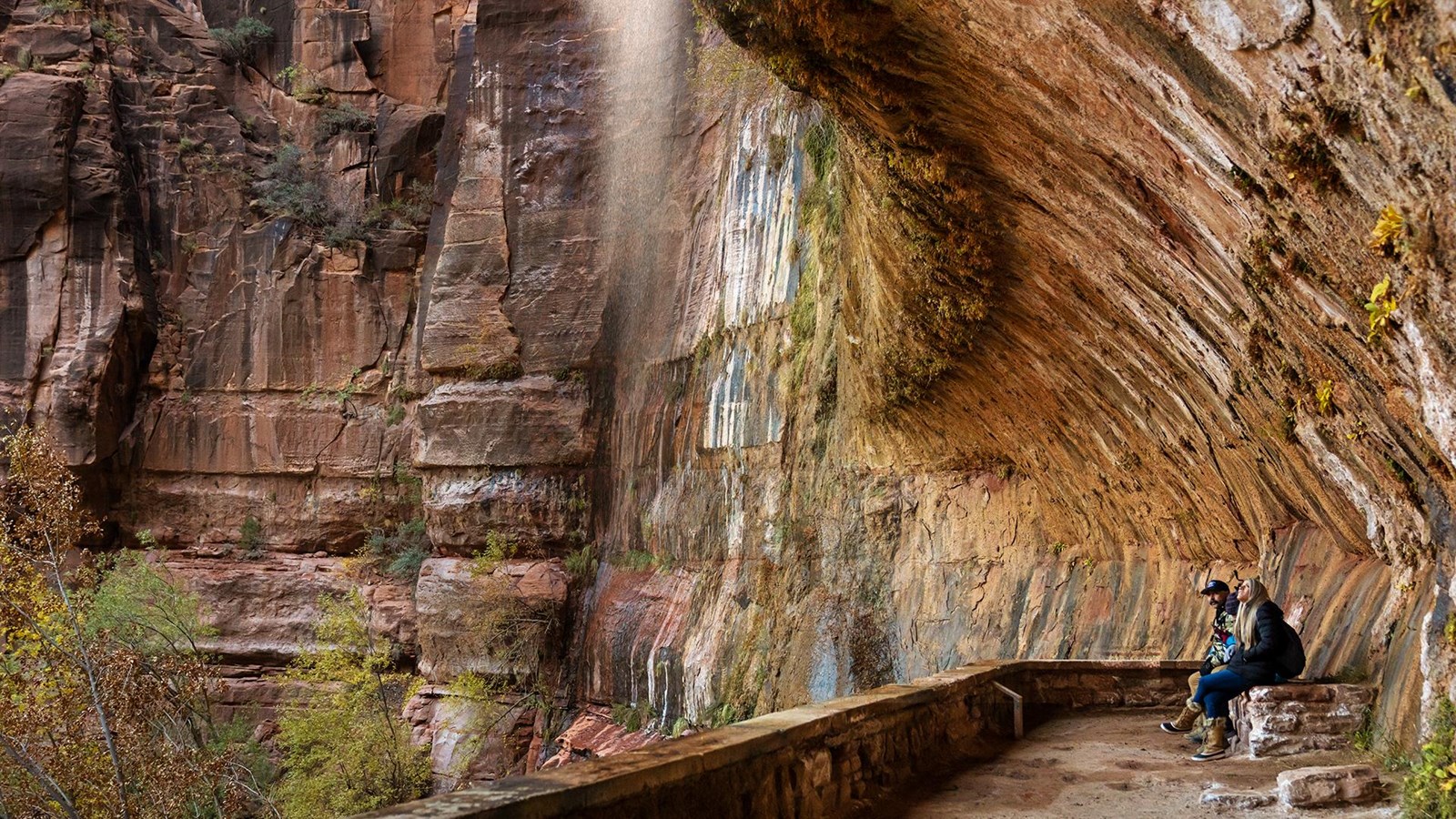 Two people sit in a rock alcove, with water dripping off the overhang in front of them.