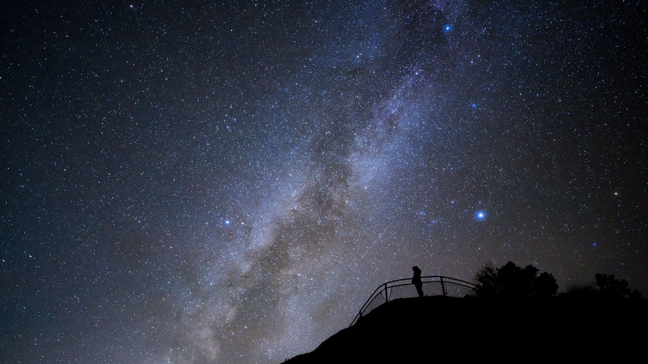 A silhouetted figure stands at a fenced overlook with the milky way arching above