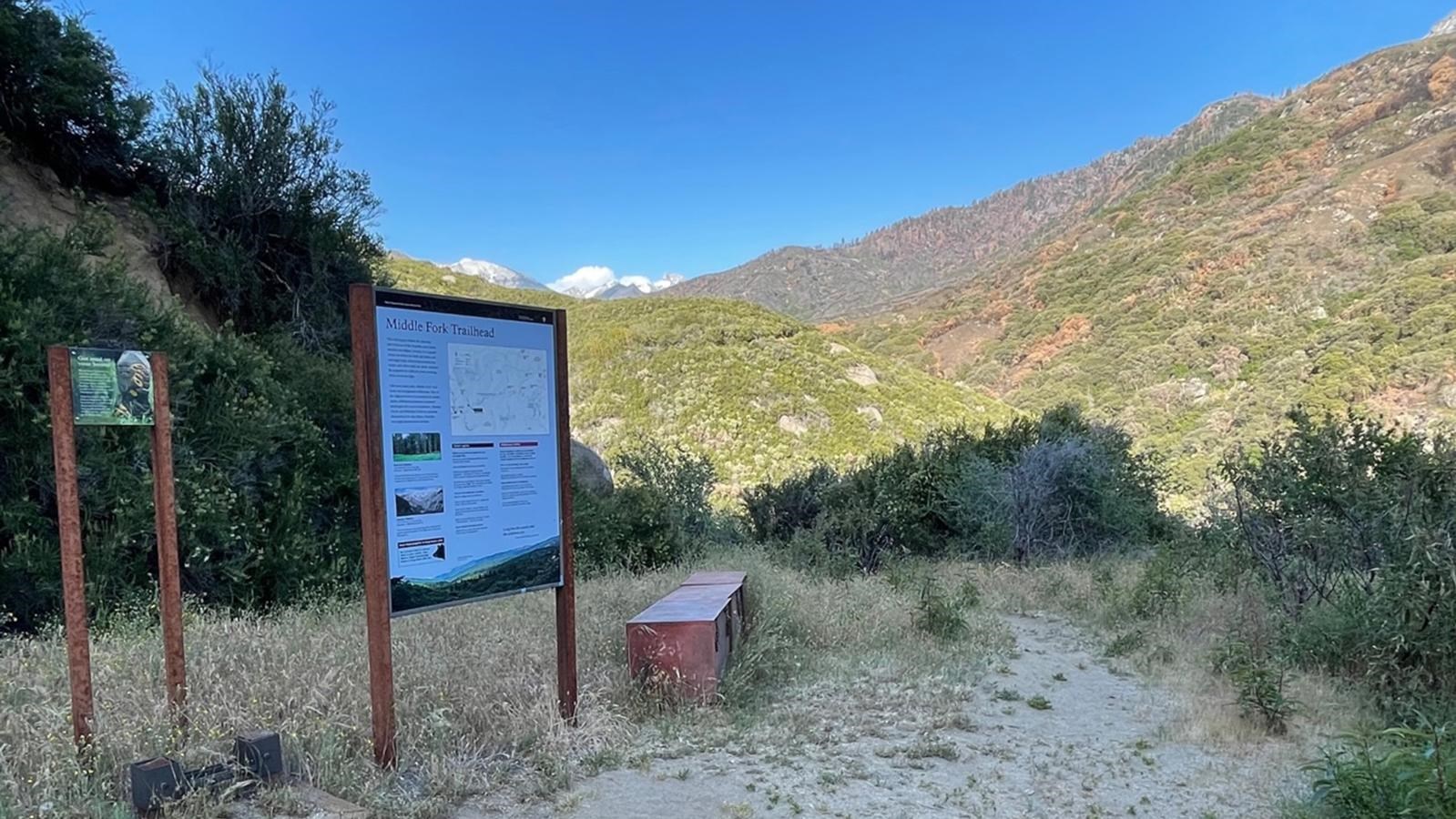 A trailhead sign and bench are located at the beginning of a dirt trail with a hill in background.