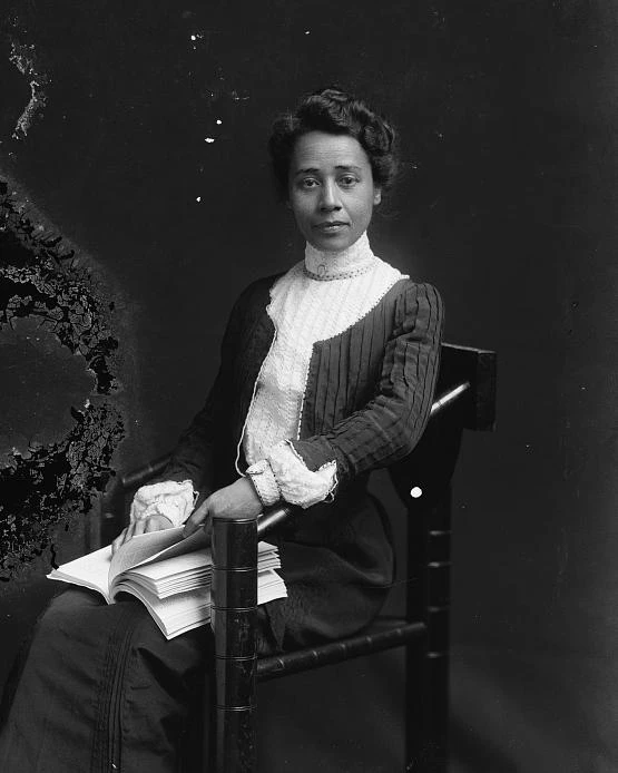 A woman sits in a chair wearing a long dress with a book on her lap, looking at the viewer