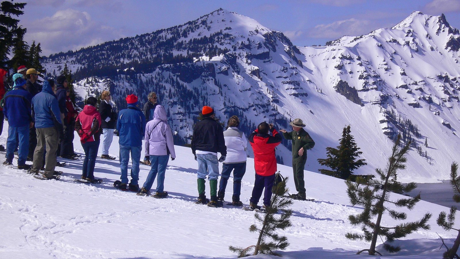 On a winter day, a ranger is encircled by visitors wearing snowshoes Crater Lake in the background